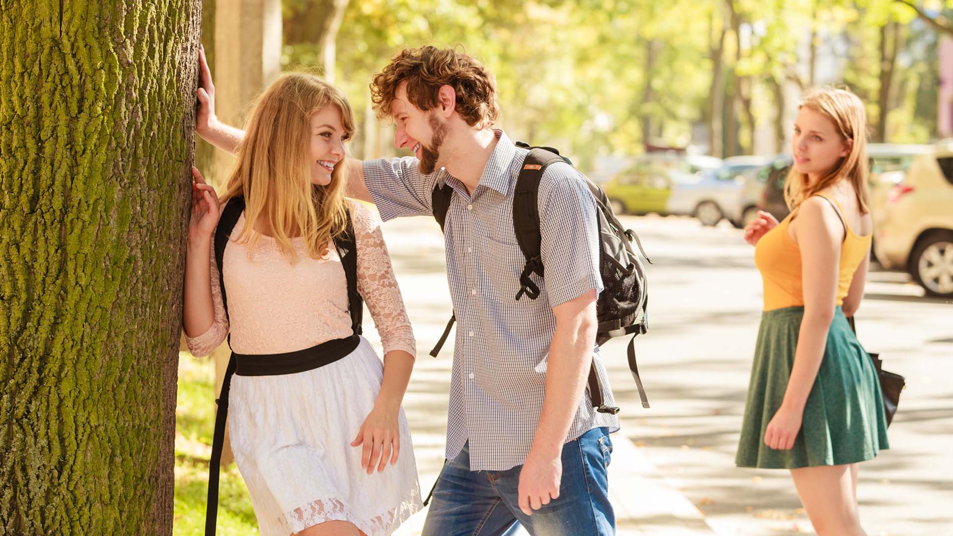 Man pinning a blonde woman against a tree and smiling at each other while another blonde woman stares at them.